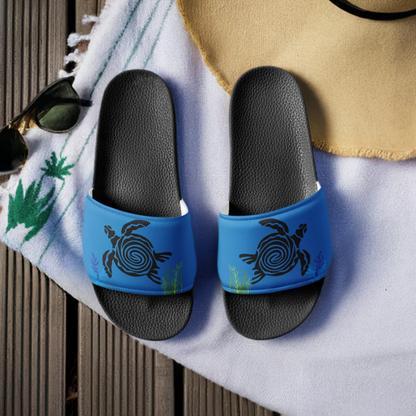 Blue slides/flip flops with a black turtle design on a deck with a beach towel and sunglasses. The slides have a black sole.