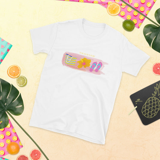 White crew neck t-shirt with pink watercolor design across the chest and has a yellow and blue beach tote, orange and pink hibiscus flower and pink flip flops with blue straps. Above the watercolor has the text Tuscan Sun Surf.